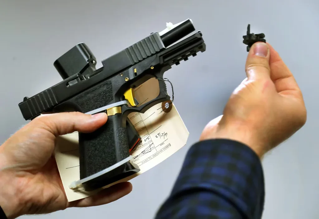 A law enforcement officer holds a Glock-style pistol bearing an evidence identification tag in one hand, and a small pistol converter device — commonly called a Glock switch — in the other, displaying both items separately against a gray background.