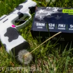 New Republic 9mm Luger 124 Grain FMJ Training Ammunition 50-Round box beside a cow-print pistol resting on grass